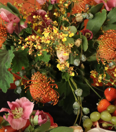 A colorful floral arrangement with pink, yellow, and orange flowers, surrounded by green leaves. In the foreground, there are fresh strawberries and green grapes on a black surface.