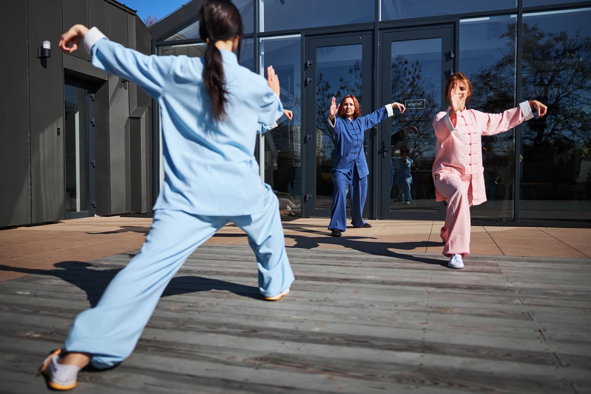 Three people in traditional martial arts uniforms practice Tai Chi outdoors on a wooden deck in front of a modern glass building under a clear sky.