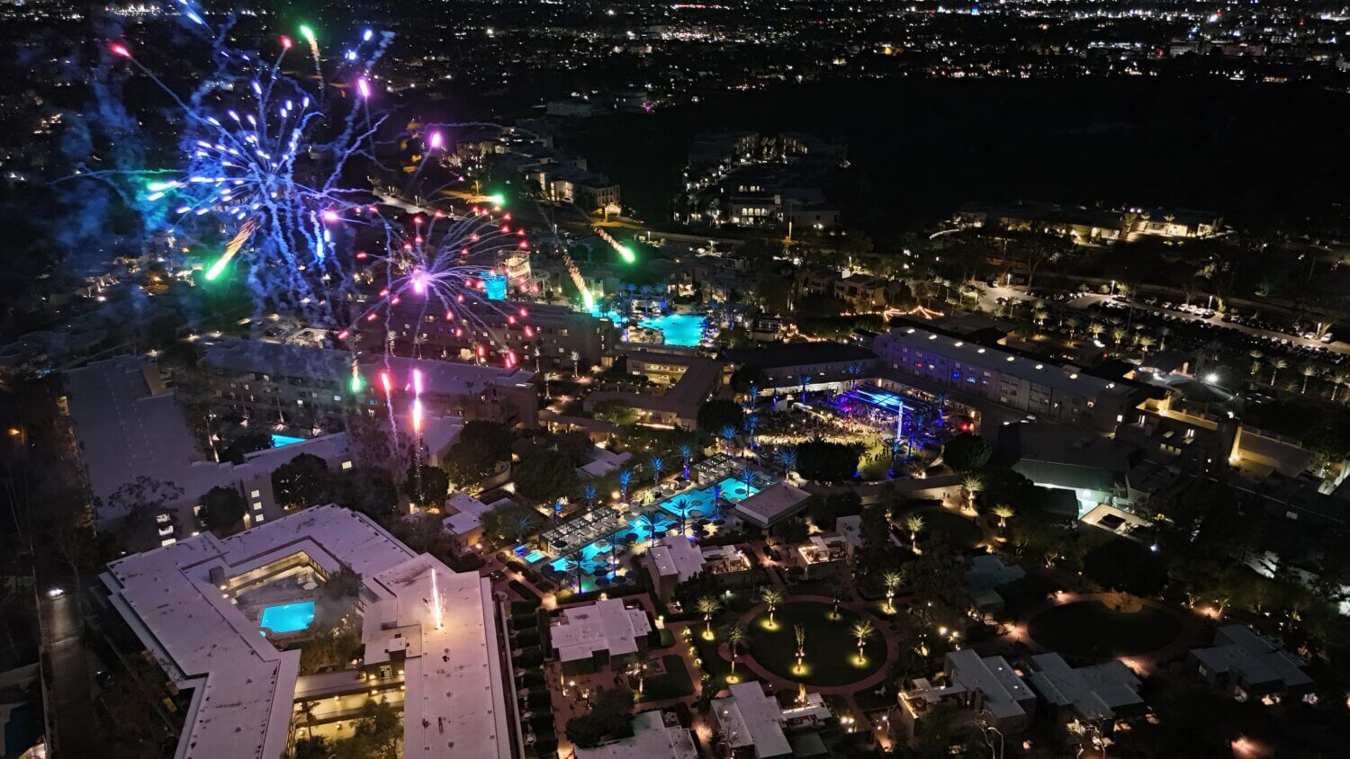 Aerial night view of a brightly lit Phoenix resort city during the Fourth of July, with colorful fireworks bursting in the sky, illuminating pools, buildings, and surrounding areas against a dark backdrop.