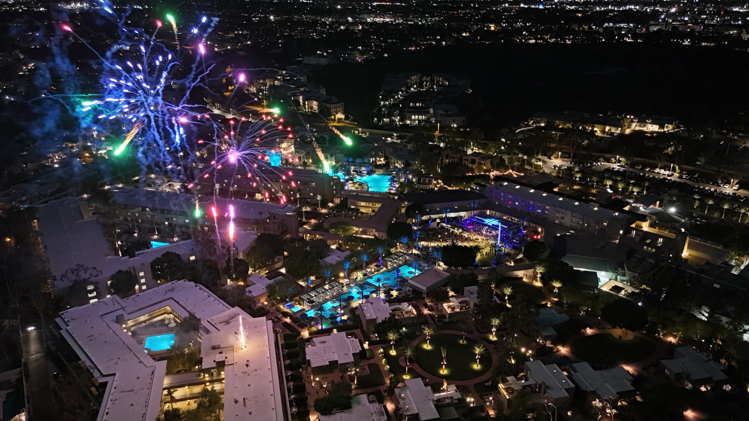 Aerial night view of a brightly lit Phoenix resort city during the Fourth of July, with colorful fireworks bursting in the sky, illuminating pools, buildings, and surrounding areas against a dark backdrop.