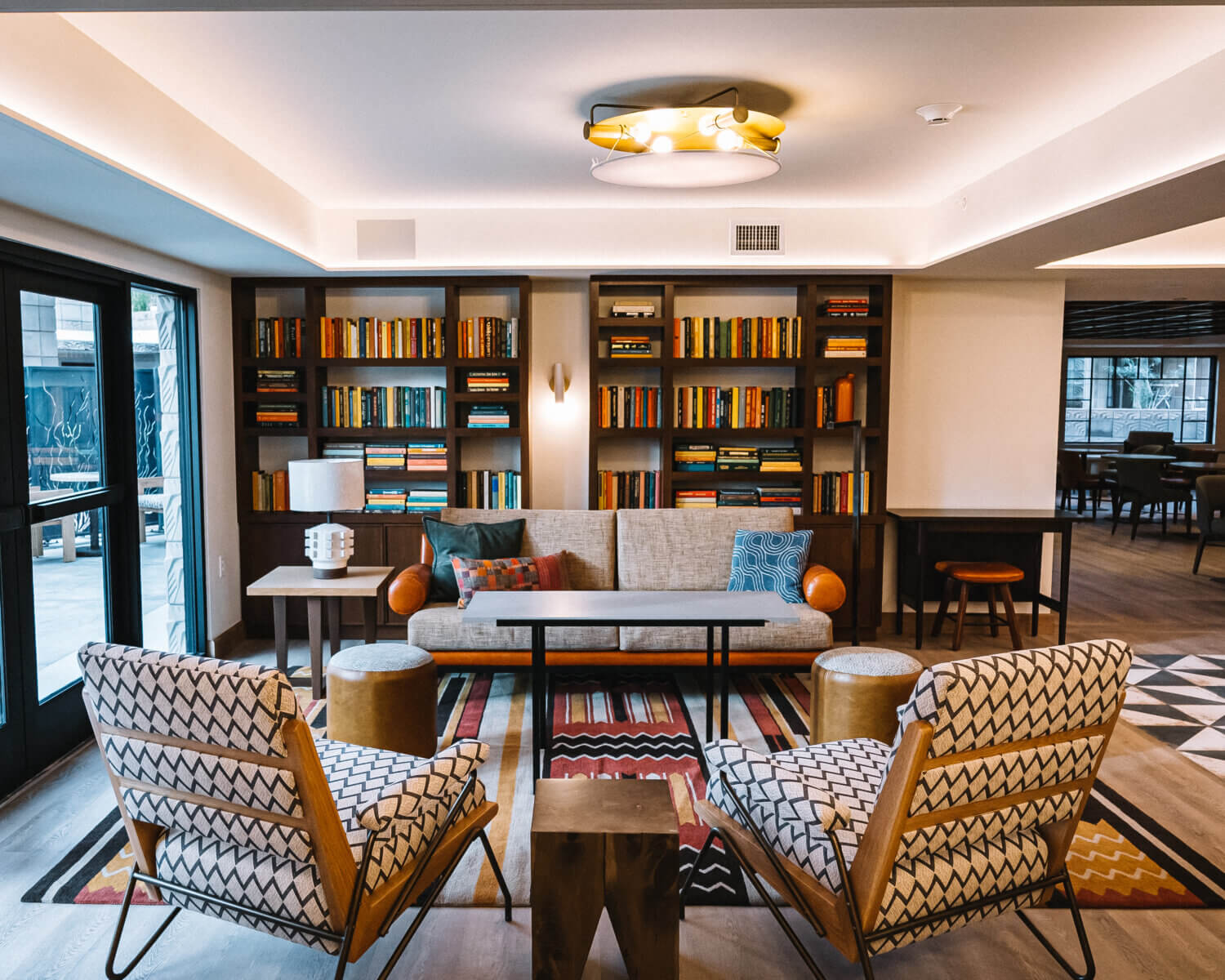 A modern lounge with a gray sofa, patterned armchairs, bookshelves of colorful books, a geometric rug, and warm lighting creates a cozy atmosphere—perfect for relaxing or enjoying the exclusive Arizona Resident Offer.