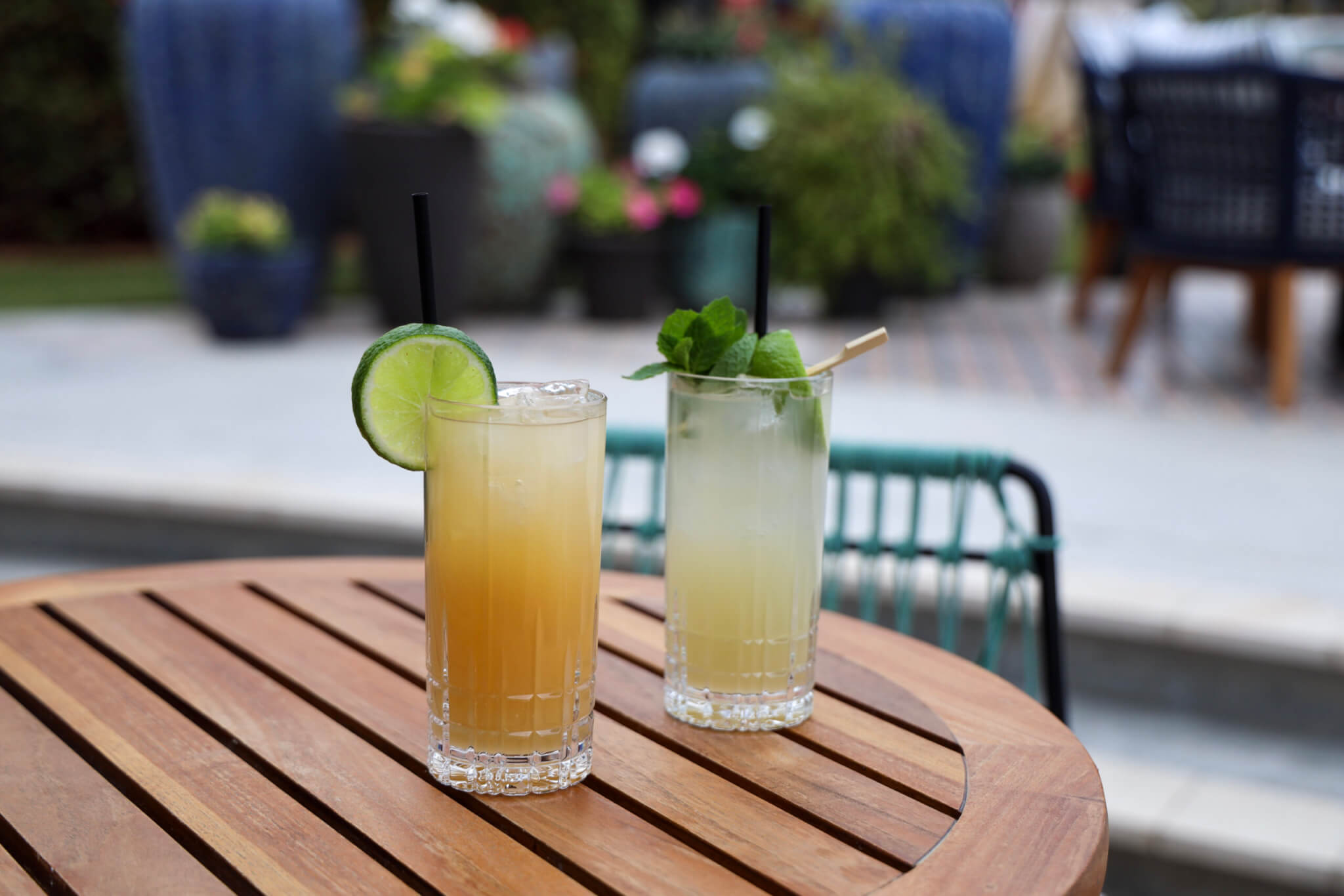 Two tall glasses of cocktails on a wooden table outdoors; one drink is orange with a lime slice, the other is light yellow with mint garnish. Blurred garden and patio furniture in the background.