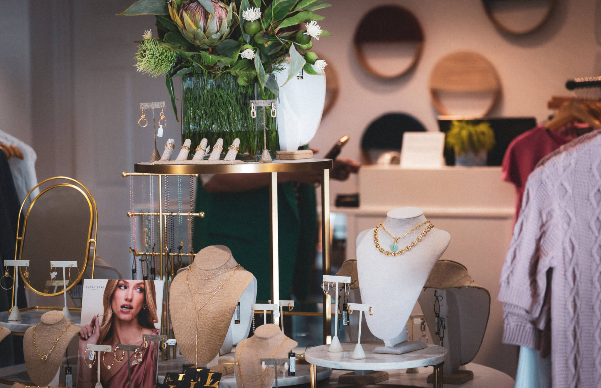 A retail jewelry store display features necklaces, rings, and earrings on white stands, with a bouquet of flowers and a card showing a model wearing jewelry. Clothing and a checkout counter are visible in the background.