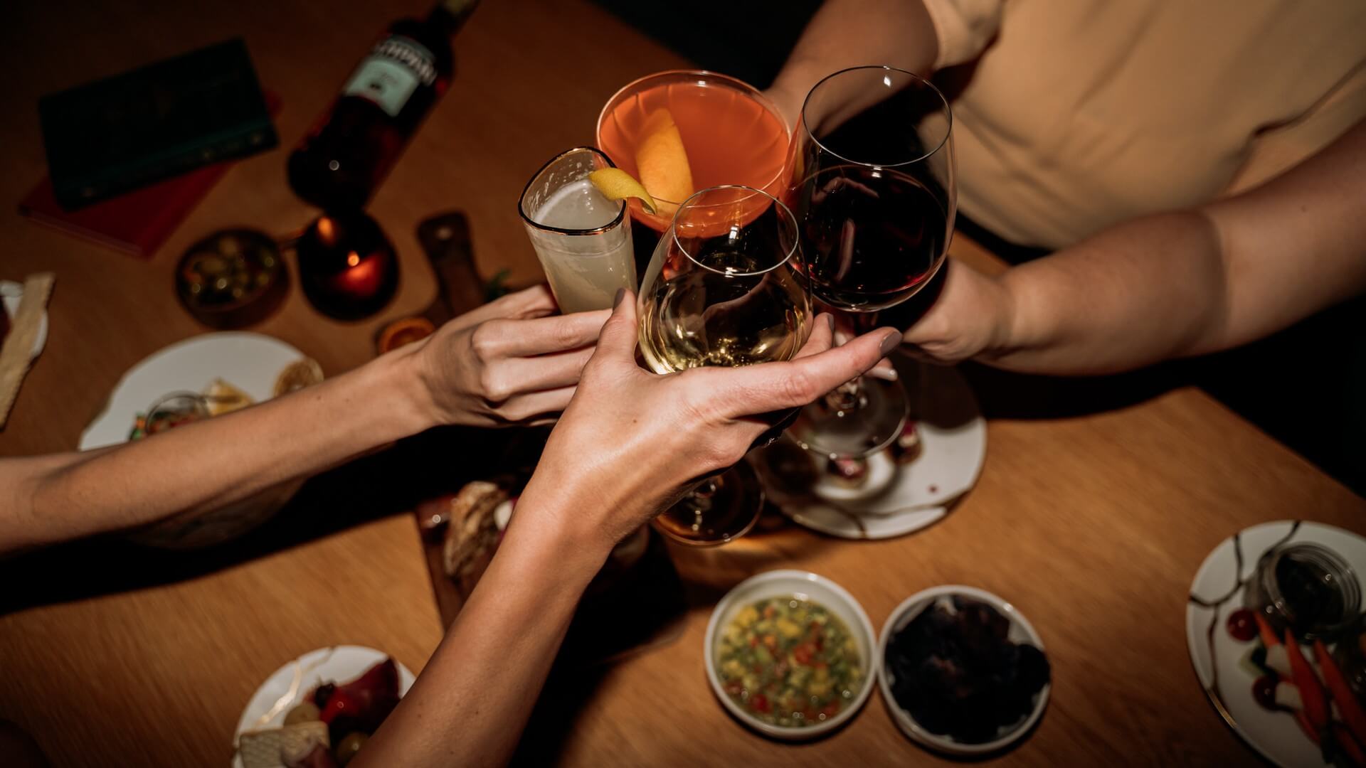 Four people clink glasses of wine and cocktails over a wooden table with plates of food, capturing a celebratory toast in a warm, festive atmosphere.