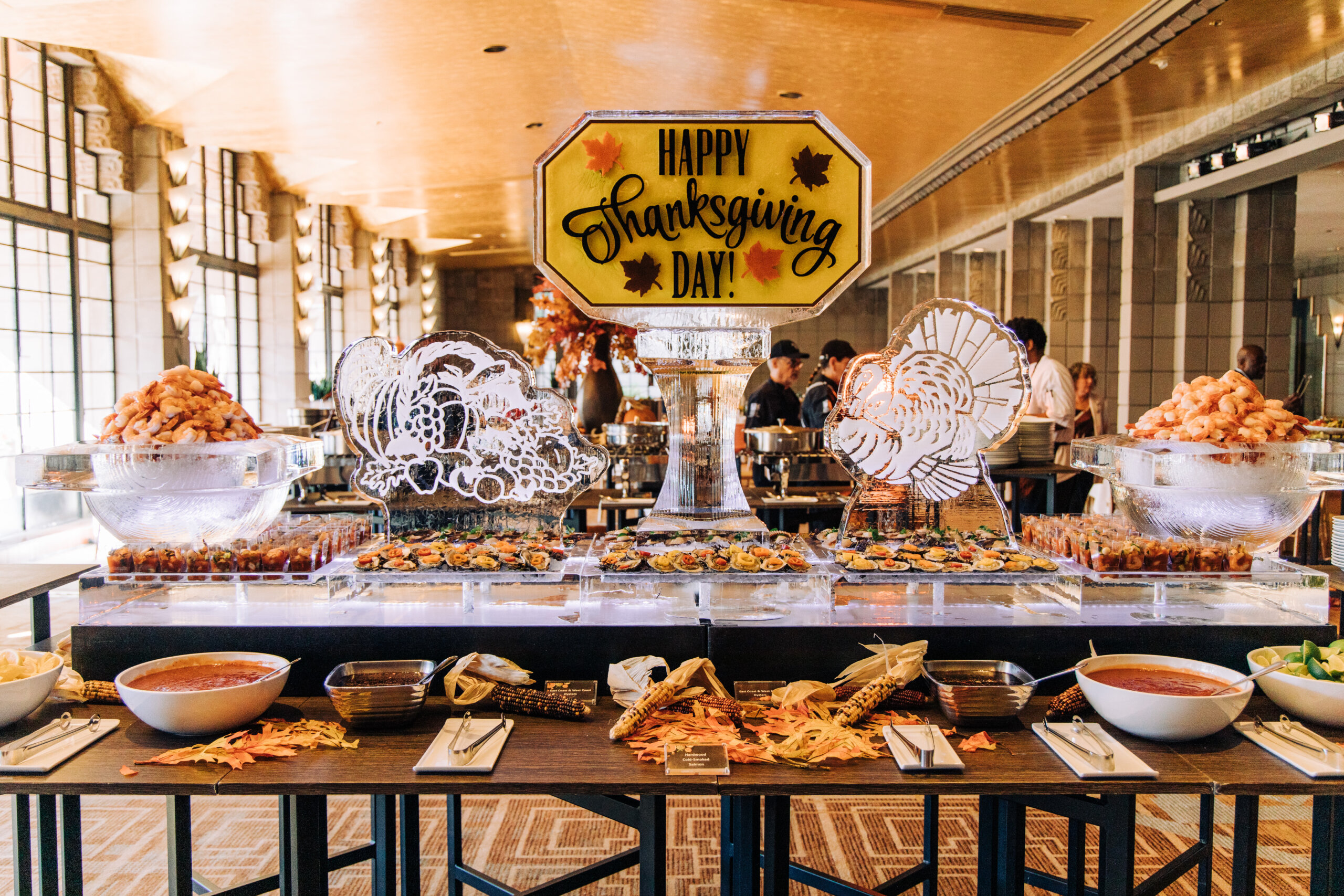 A Thanksgiving buffet with an ice sculpture centerpiece that says “Happy Thanksgiving Day!” Flanking the center are two more ice sculptures of turkeys, shrimp platters, seafood displays, bowls of cocktail sauce, and decorative autumn leaves.