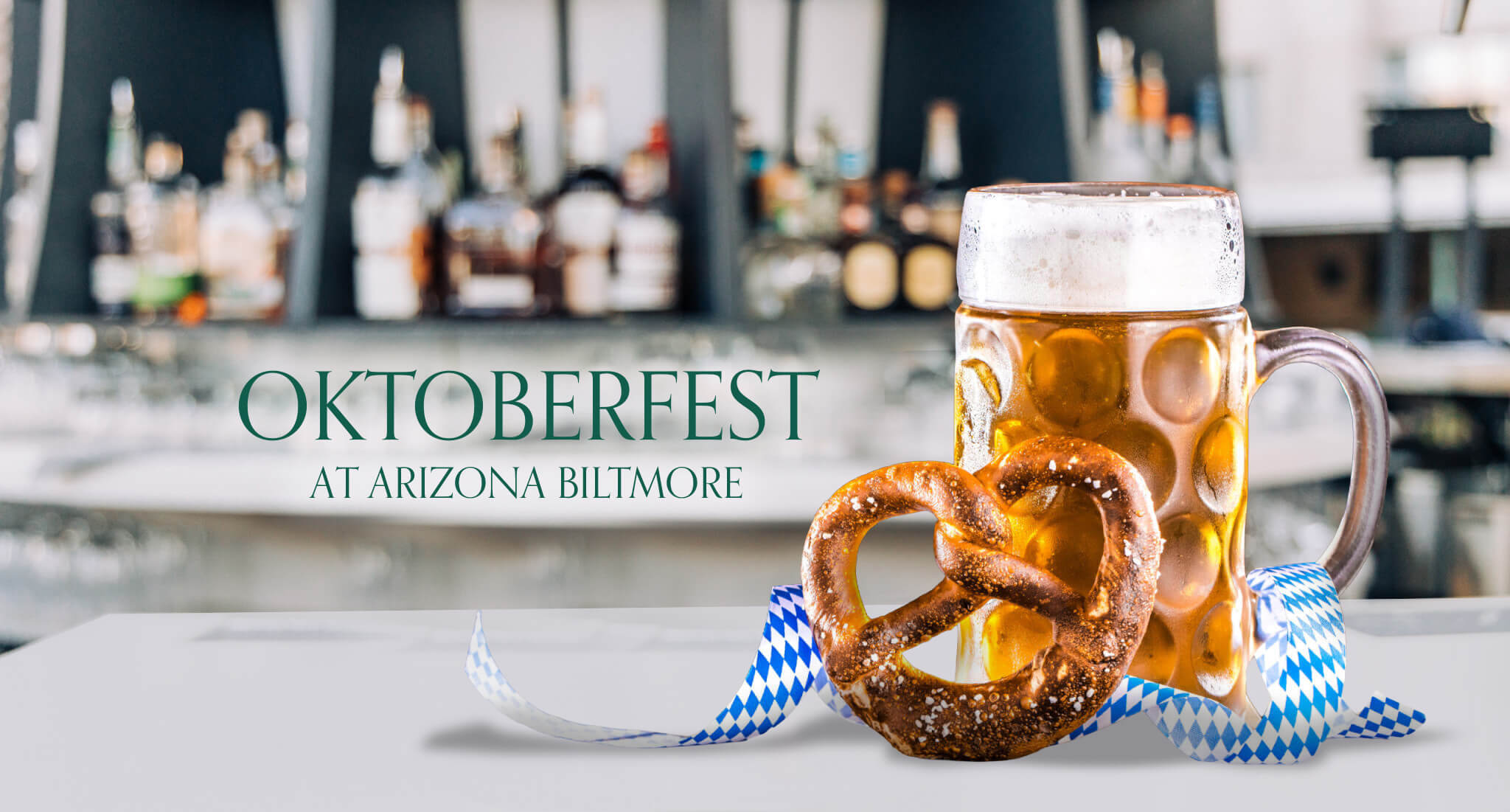 A large beer mug and a salted pretzel sit on a table with blue and white ribbon. The background shows blurred liquor bottles. Text reads “OKTOBERFEST AT ARIZONA BILTMORE.”.