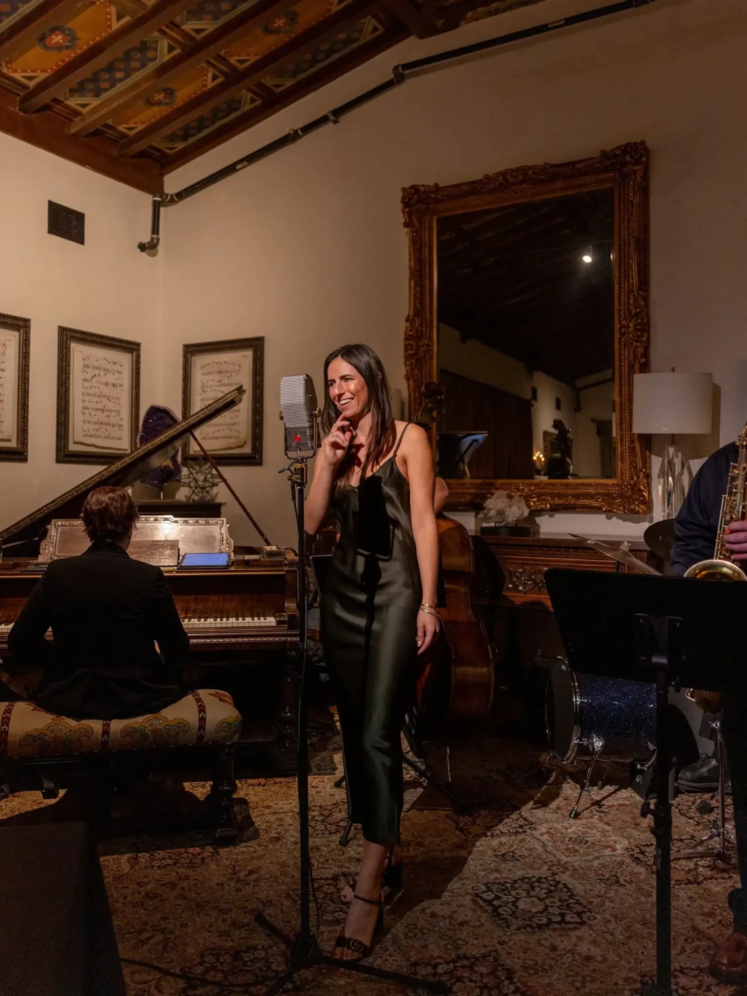 A woman in a dark dress sings into a microphone, accompanied by a pianist and a saxophonist in a dimly lit room.