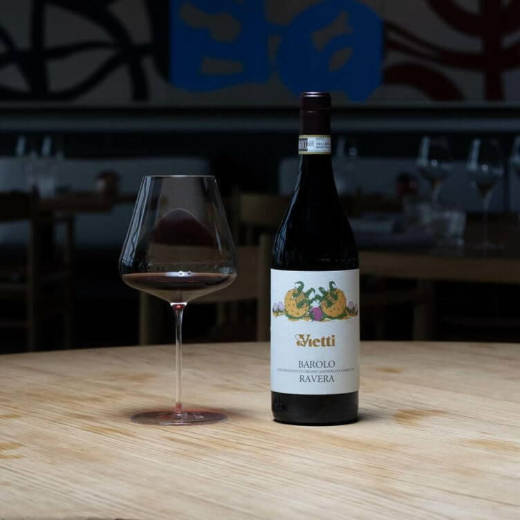 A bottle of Vietti Barolo Ravera wine next to a glass filled with red wine on a wooden table.