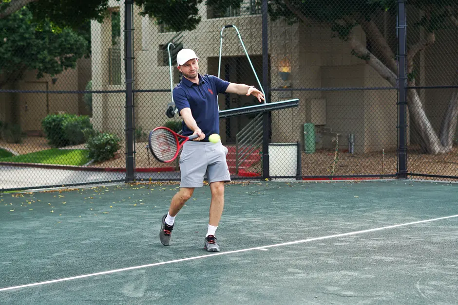 A man in a navy polo shirt and grey shorts hits a tennis ball on a court.